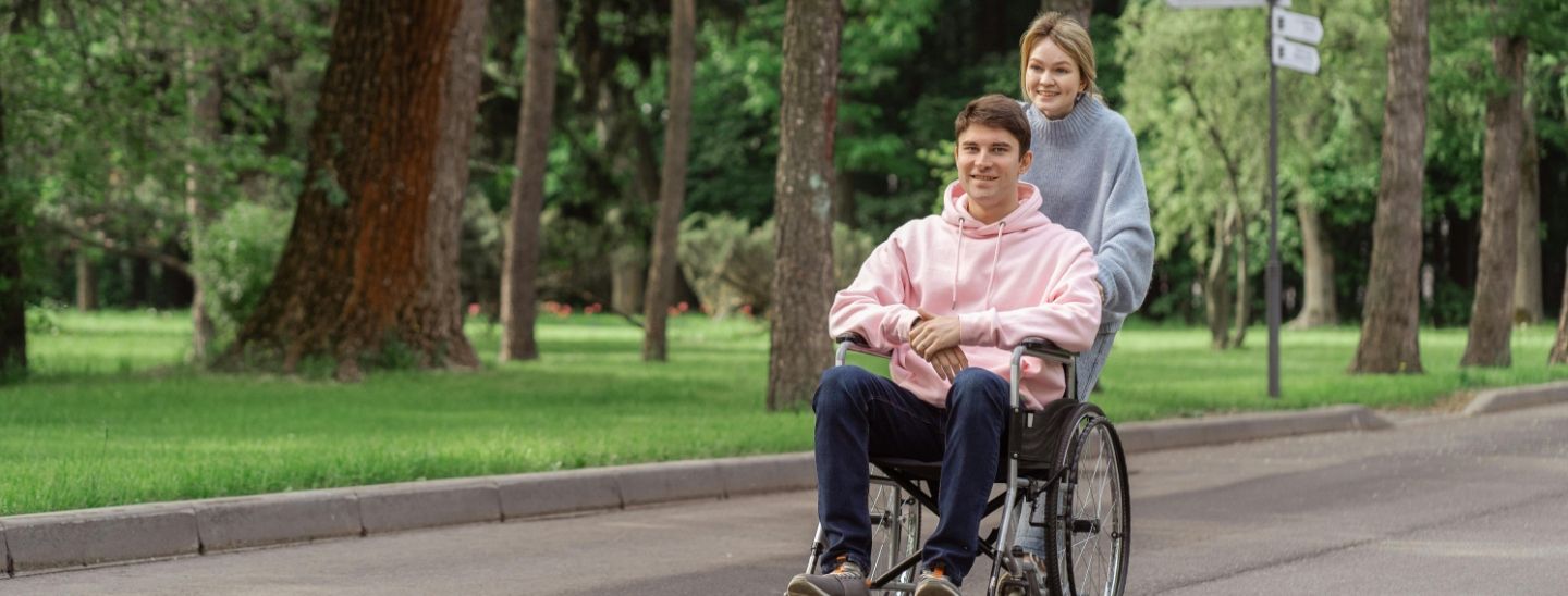A man in a wheelchair being pushed along a paved park path by a female companion on a sunny day.