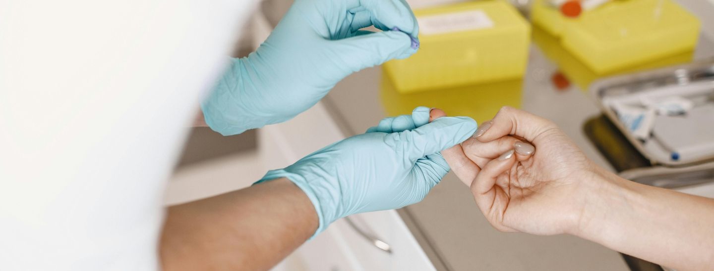 A healthcare professional in blue gloves performing a finger prick blood test on a patient in a clinical setting.