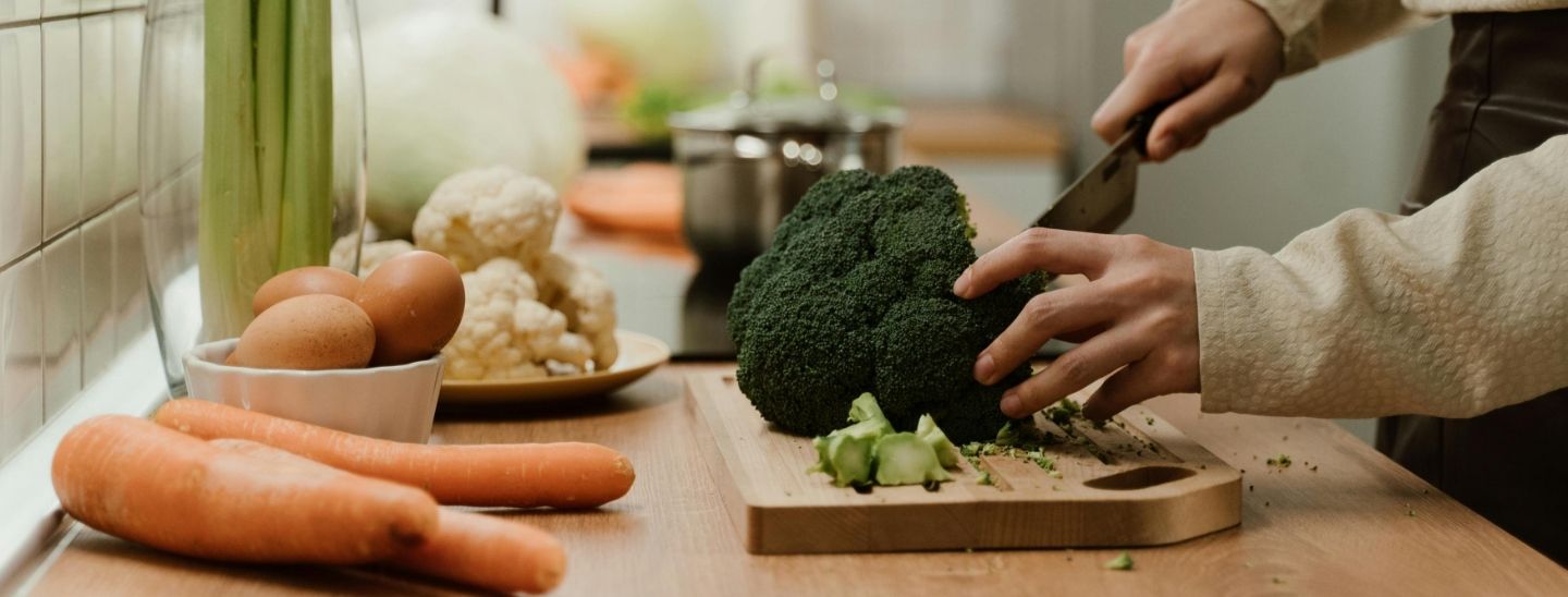 Person’s hands using a chef's knife to chop a head of fresh broccoli on a wooden cutting board.
