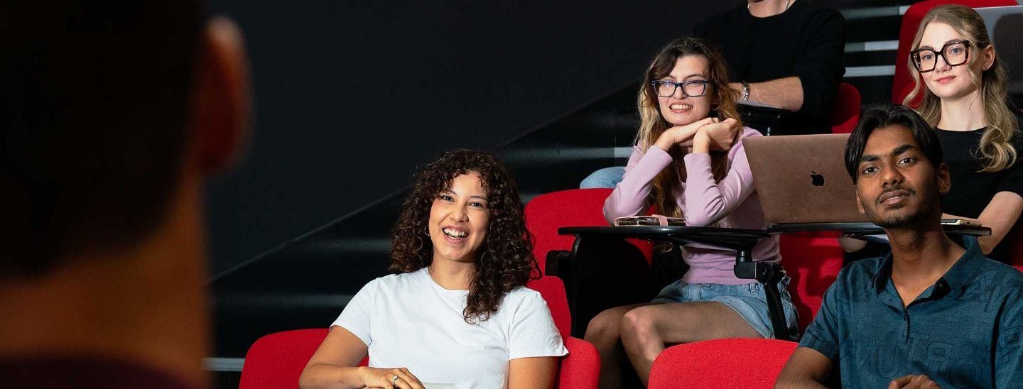 ECC students sitting in red theater-style seats in a lecture hall, some using laptops and smiling toward the camera.