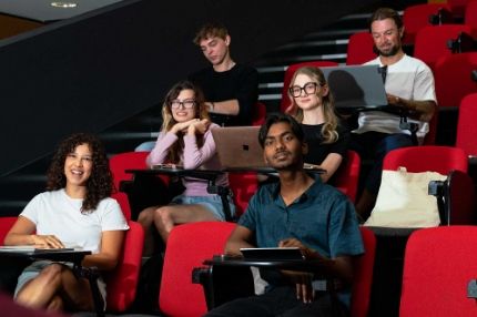 ECC students sitting in red theater-style seats in a lecture hall, some using laptops and smiling toward the camera.