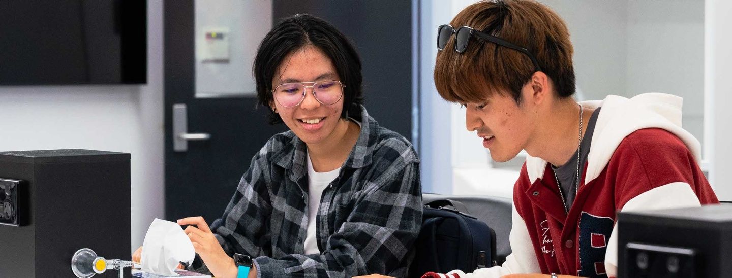 Two students sitting at a white table in a classroom setting, looking at a notebook and discussing their work.