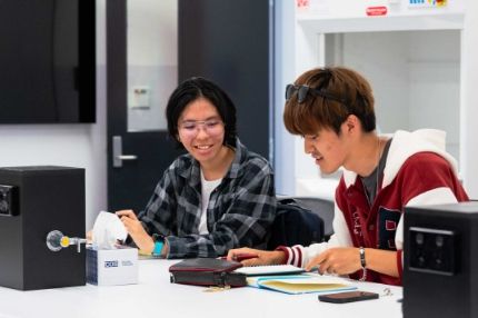 Two students sitting at a white table in a classroom setting, looking at a notebook and discussing their work.