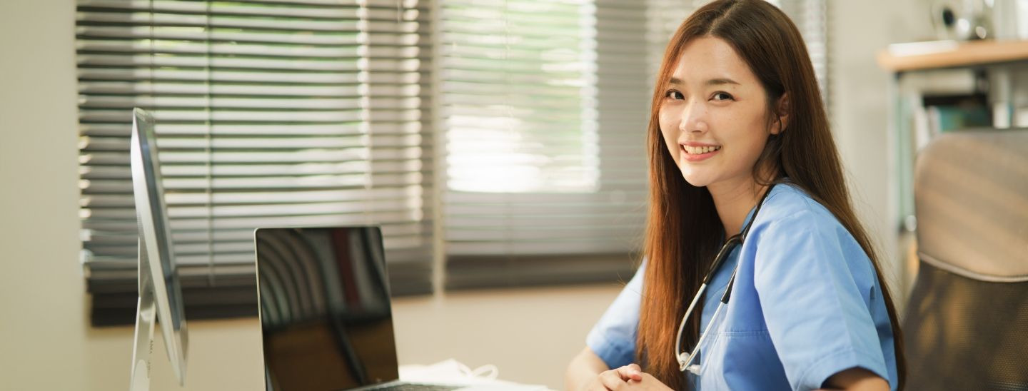 Female nurse in blue scrubs with a stethoscope around her neck, sitting at a desk with a laptop and monitor in a bright office with window blinds.