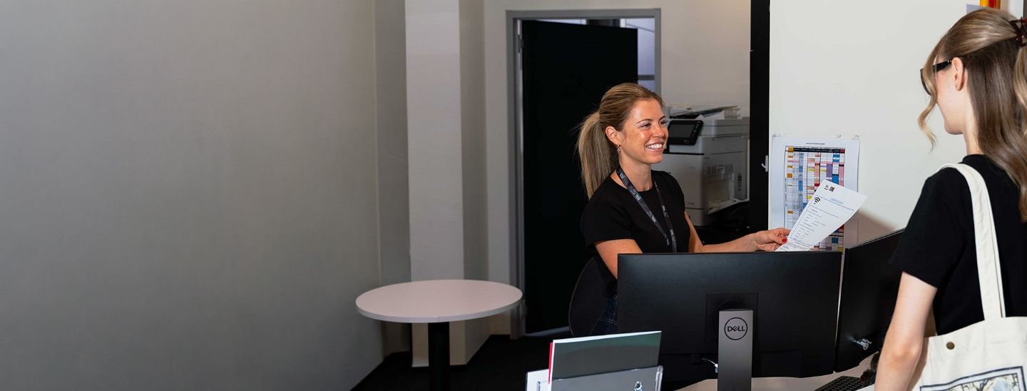 College receptionist assisting a student with paperwork at a front desk.