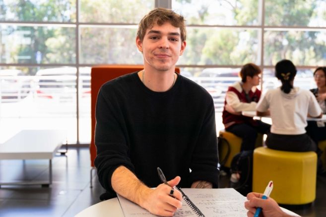 Student sitting at a table writing in a notebook and looking up