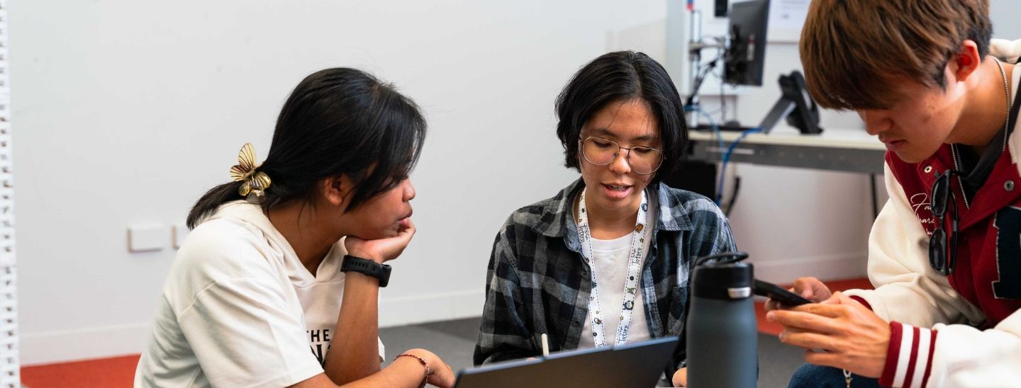 Students sitting at a white table in a classroom setting, looking at a notebook and discussing their work.