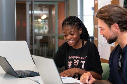 Female student looking at a laptop during a collaboration session with a mentor in a modern office or educational setting.
