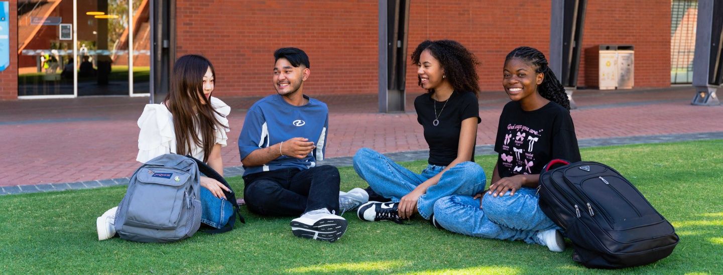 ECC students sitting on a green lawn in front of a brick campus building, talking and smiling with their backpacks beside them.