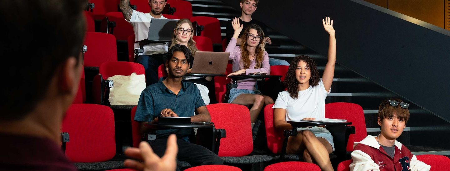ECC students sitting in red theater-style seats in a lecture hall, some using laptops and smiling toward the camera.