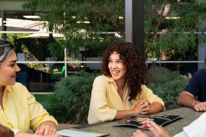 Students laughing and discussing a project while sitting at an outdoor table with notebooks.