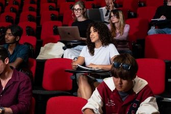 ECC students sitting in red theater-style seats in a lecture hall, some using laptops and smiling toward the camera.