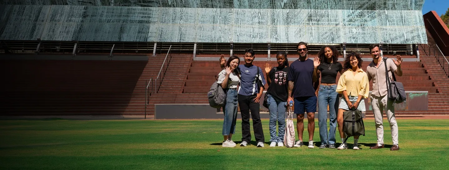 ECC students standing together on a green lawn in front of a modern campus building, waving at the camera.