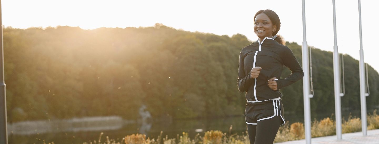 A smiling female in black athletic wear running along a lakeside path during a golden hour sunset with trees in the background.