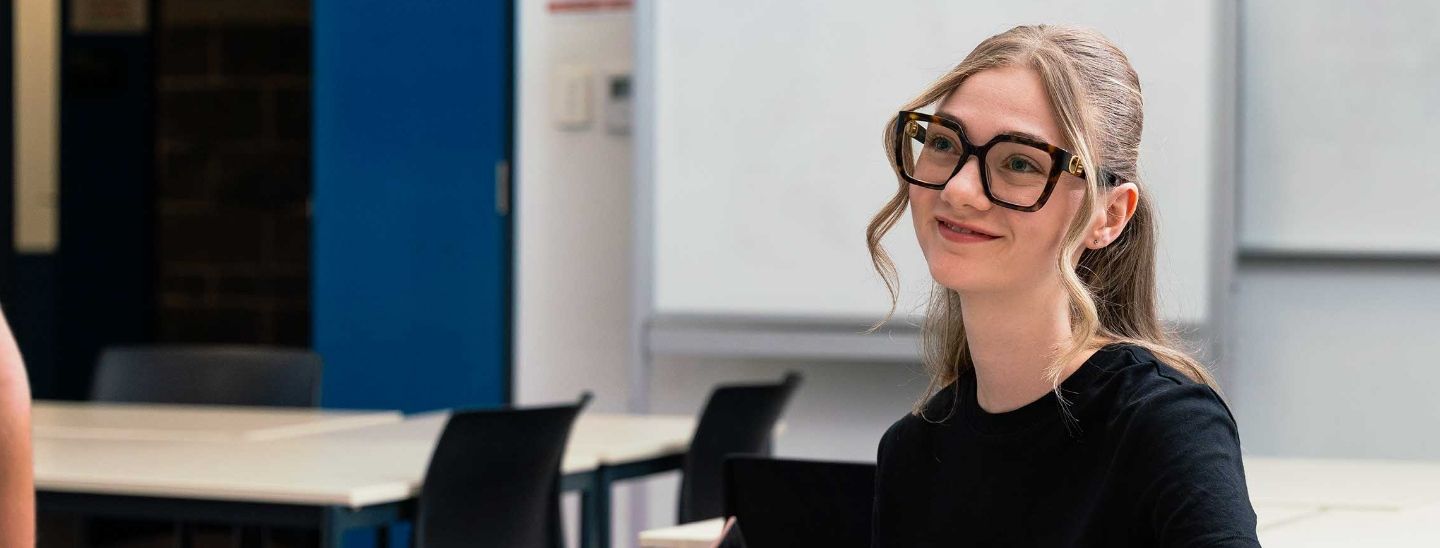 Female student wearing stylish glasses in a classroom with whiteboards in the background.