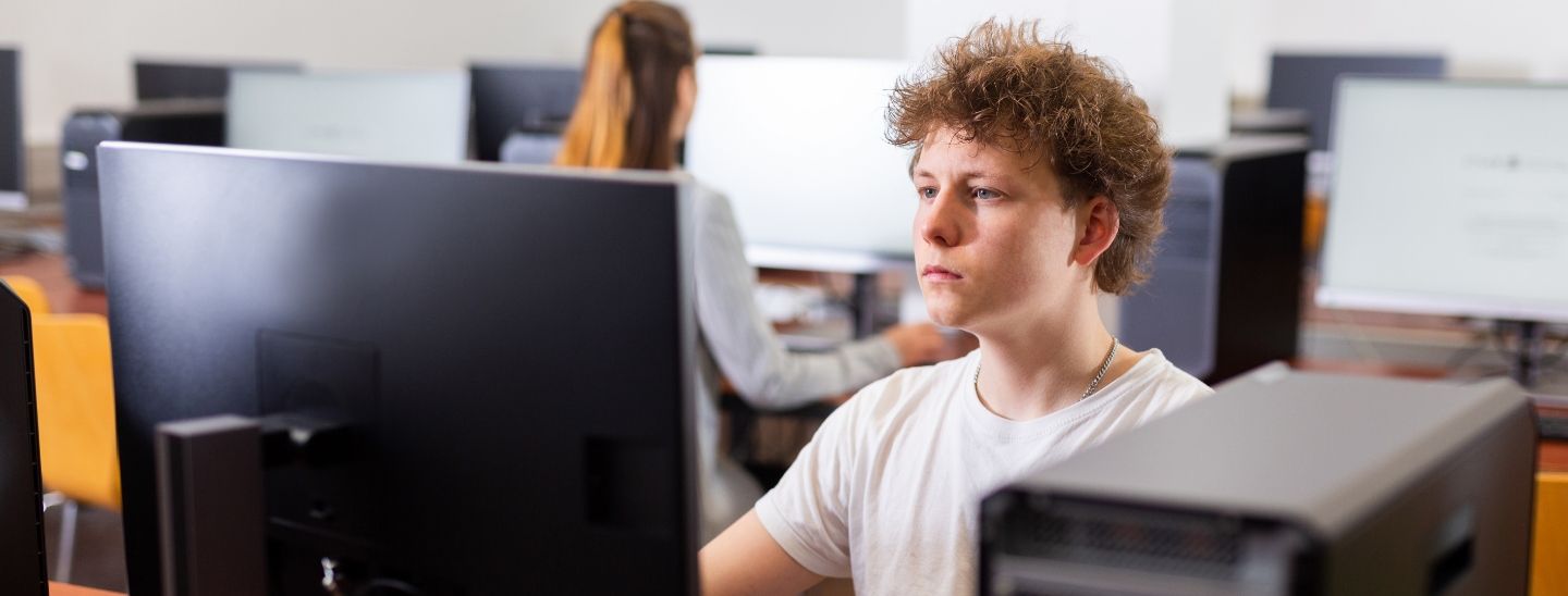 Male student working intently at a desktop computer in a modern IT lab with other students in the background.