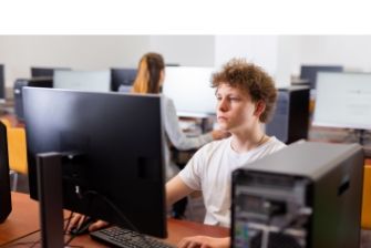 Male student working intently at a desktop computer in a modern IT lab with other students in the background.