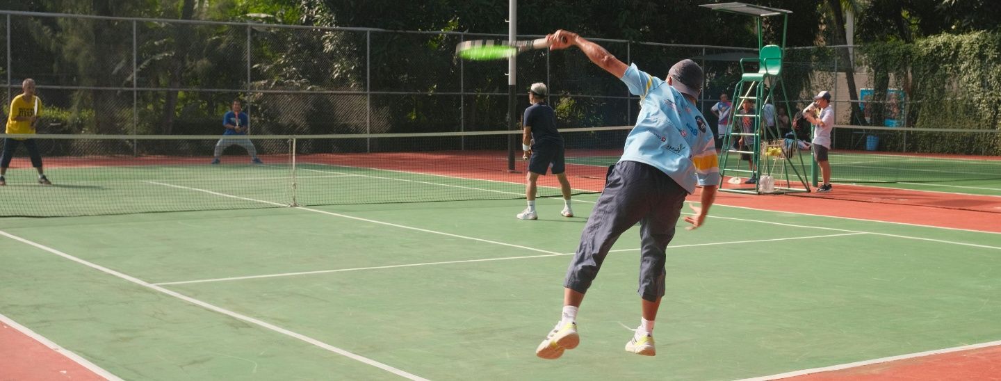An action shot of a tennis player mid-serve on an outdoor court surrounded by trees.