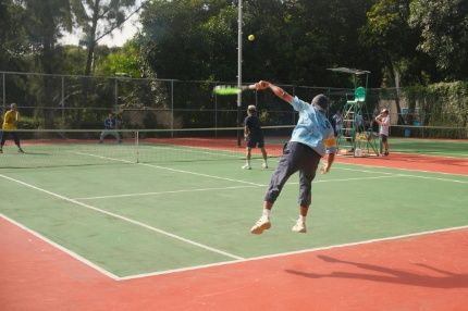 An action shot of a tennis player mid-serve on an outdoor court surrounded by trees.