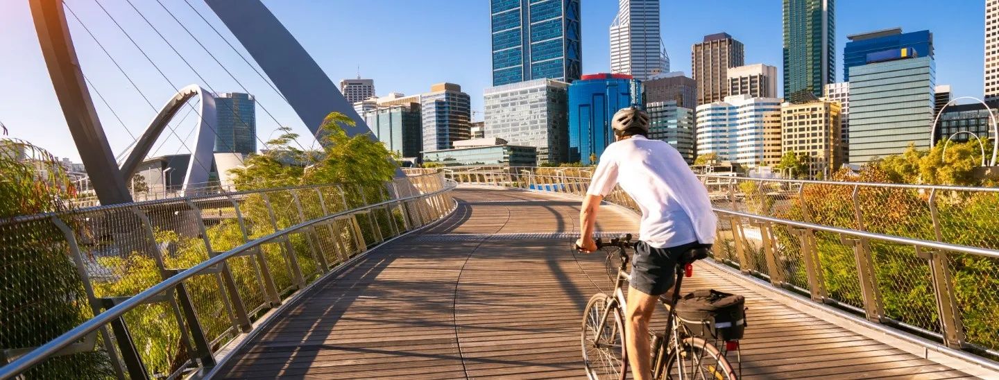 Man cycling on a bridge overlooking the Perth city skyline during a sunny day.