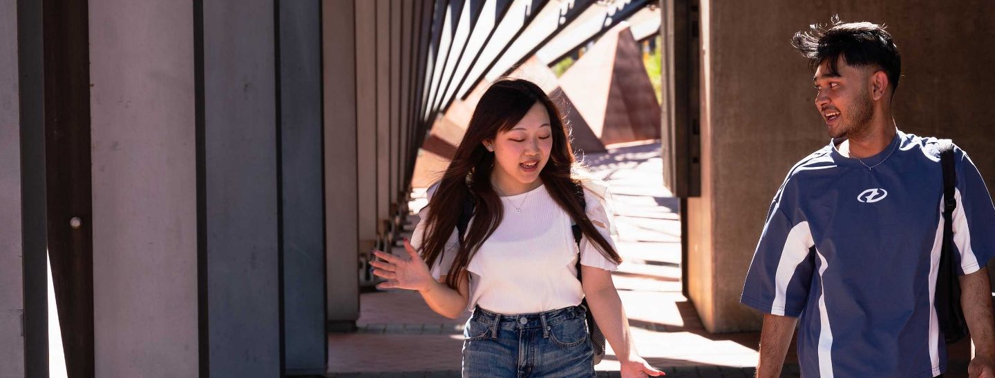 Two students talking and walking through a modern outdoor hallway on a sunny day.