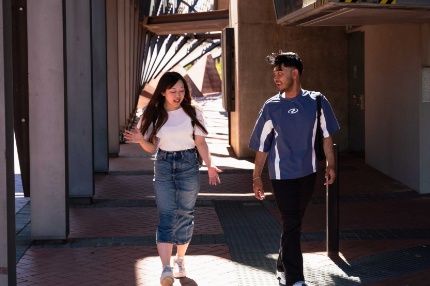 Two students talking and walking through a modern outdoor hallway on a sunny day.