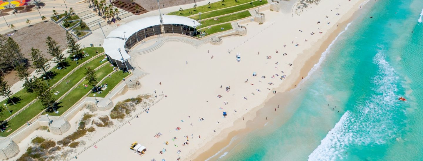 High-angle view of people relaxing on a sandy beach next to a coastal park and amphitheater.