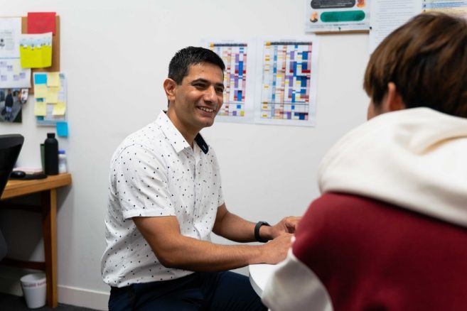 Male teacher in a white short-sleeved button-down shirt sits at a desk, engaging in a supportive conversation with a student.